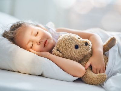 Adorable little child is sleeping in the bed with her toy. The girl is hugging the teddy bear.