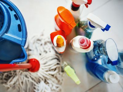 Closeup top view of unrecognizable home cleaning products with blue bucket and a mop on the side. All products placed on white tiled bathroom floor.